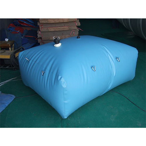 A blue, rectangular inflatable water bladder with a white valve and black cap on top, featuring metal loops on the sides, rests on a green surface with stacked materials and a blue tarp in the background.