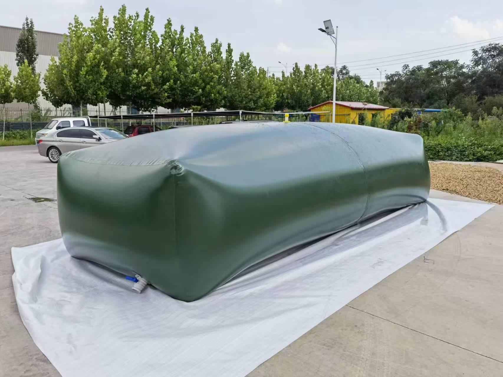 A large deep green inflatable water bladder filled with water, resting on a white tarp on a concrete surface, is surrounded by a parking lot with cars, trees, and a yellow building in the background under a cloudy sky.