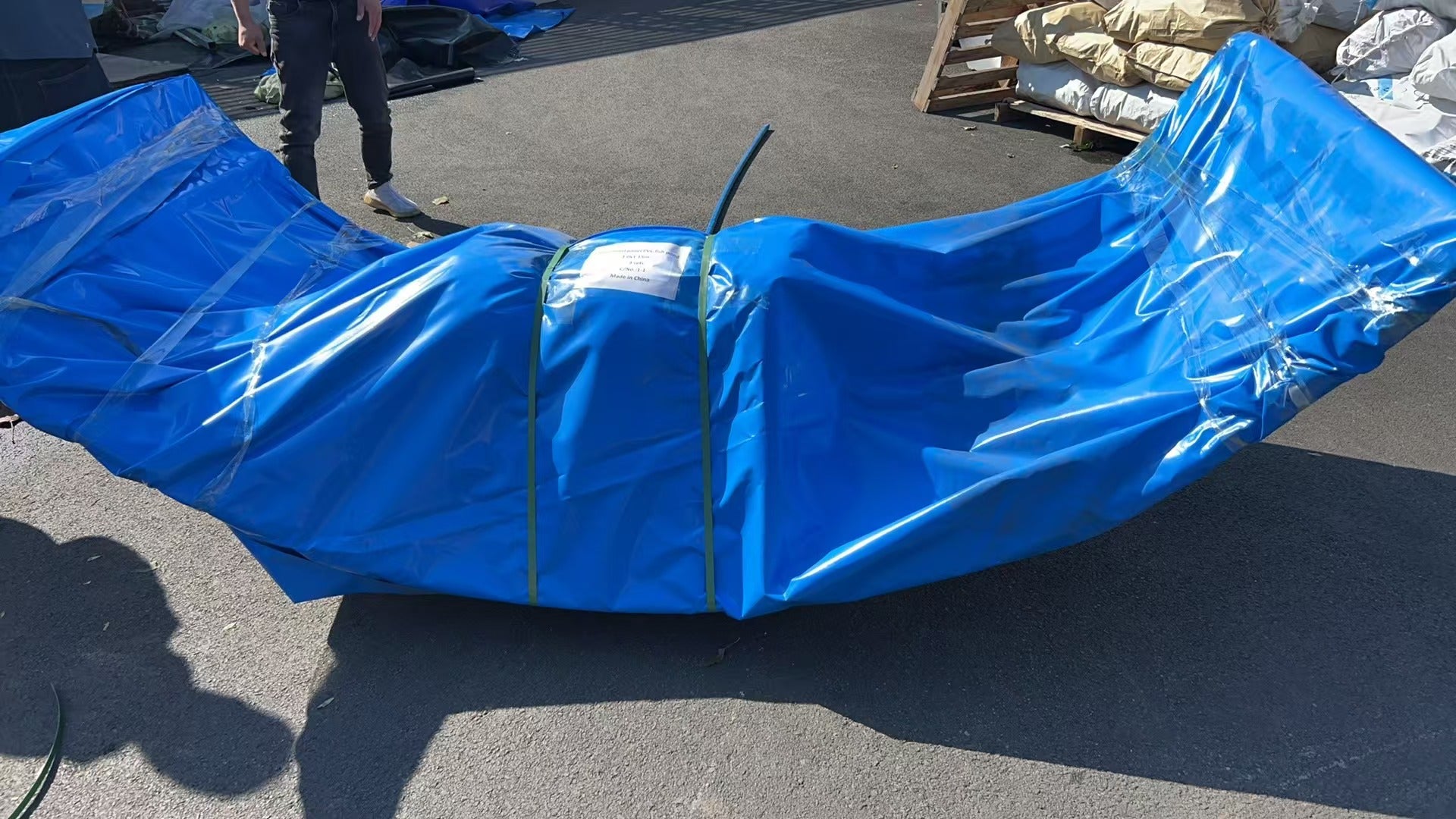 A large object wrapped in blue plastic tarp and secured with green straps is placed on the ground outdoors. A person stands nearby, casting a shadow, with stacks of materials and wooden pallets in the background