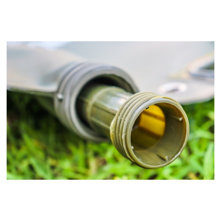 A close-up of a gray portable fuel tank, ribbed hose connected to a black, threaded cap, resting on a grassy surface. The hose has a flexible, corrugated design, and the cap is securely attached with visible threading.