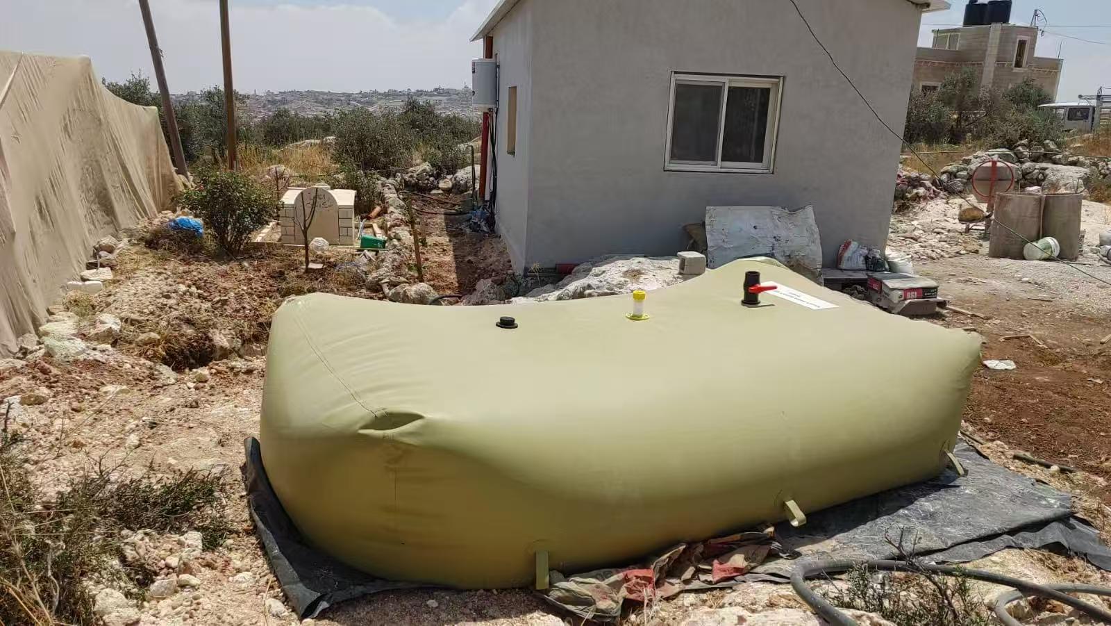 A large beige inflatable water bladder with a black cap and a red-handled valve, resting on a black tarp, is placed on a rocky terrain near a small house with a window, surrounded by scattered debris, plants, and a tent-like structure under a clear sky with distant hills visible.