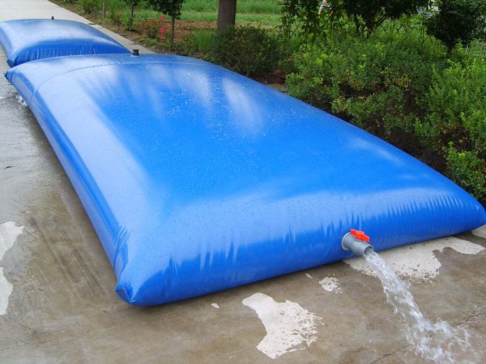 Two Large rectangular blue inflatable water storage bladder with a red valve releasing water onto a concrete surface, surrounded by greenery, ideal for emergency water storage, irrigation, or outdoor water management.