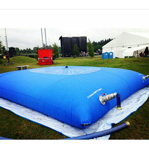 A Large blue inflatable water storage bladder with a white valve and hose, placed on a grassy field with a white protective sheet, surrounded by outdoor event equipment like tents and portable toilets, ideal for temporary water storage at events or festivals.