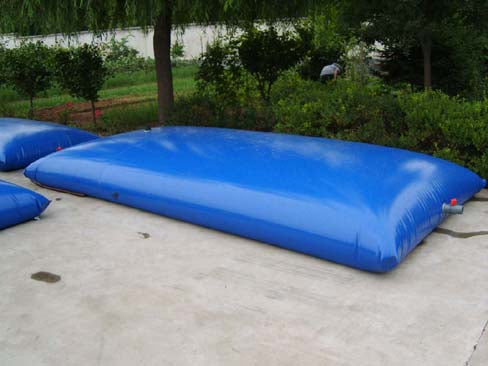A Large rectangular blue inflatable water storage bladder with a red valve and hose, placed on a concrete surface outdoors surrounded by greenery, perfect for emergency water storage or agricultural irrigation.