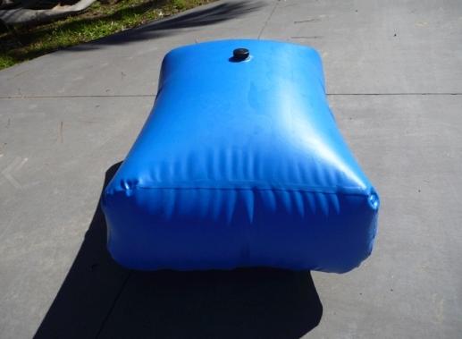 A blue inflatable water bladder with a black cap on top lies on a concrete surface, casting a shadow and surrounded by some grass in the background.
