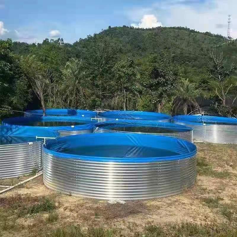 A series of large, circular fish metal tanks with blue plastic tarps covering the tops are set up outdoors on a grassy area. The tanks are surrounded by dirt and vegetation, with a forested hill in the background under a partly cloudy sky