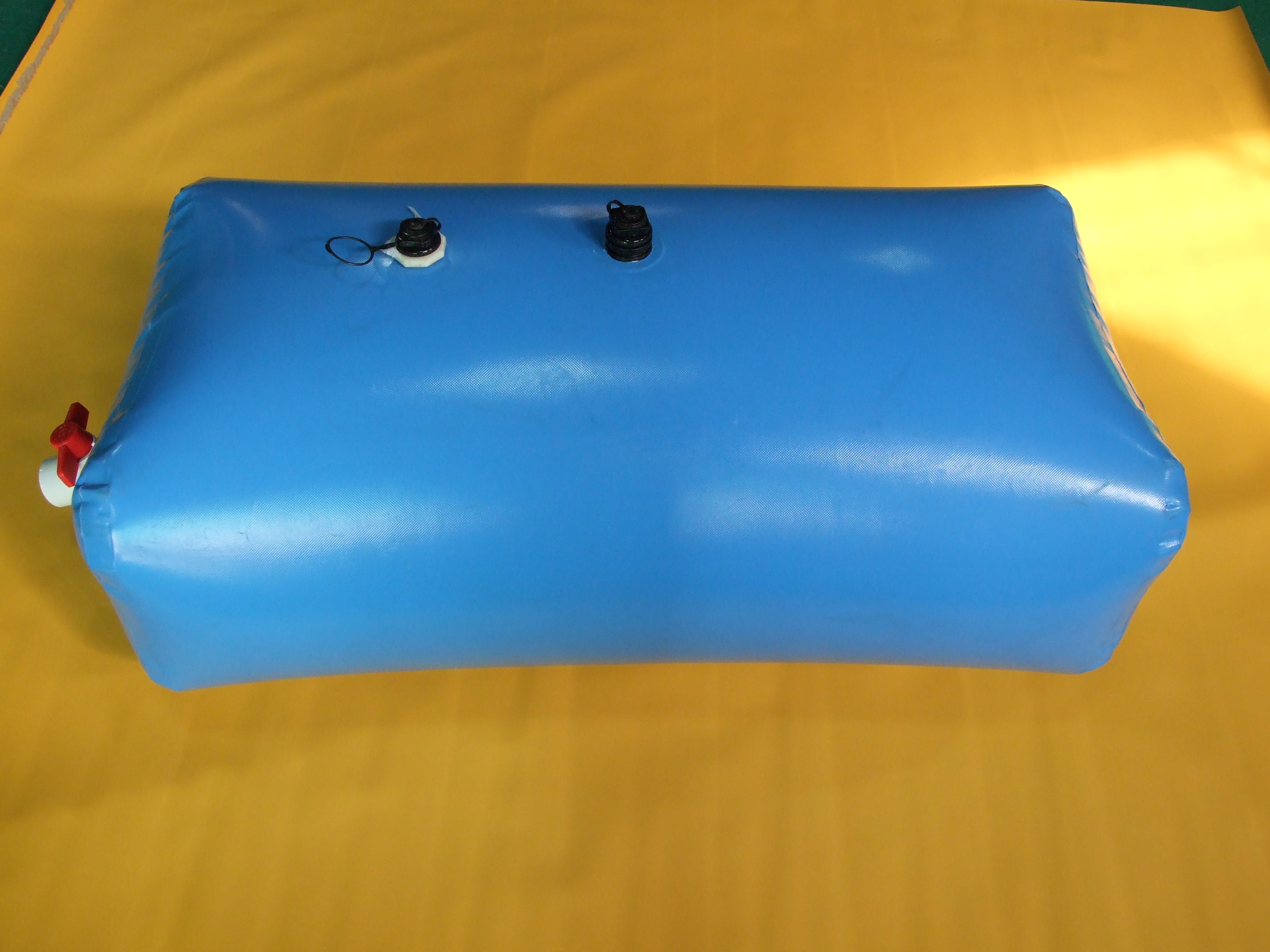 A blue, rectangular inflatable water bladder with two black caps on top and a white valve with a red cap on the side lies on a yellow surface, filled with water.