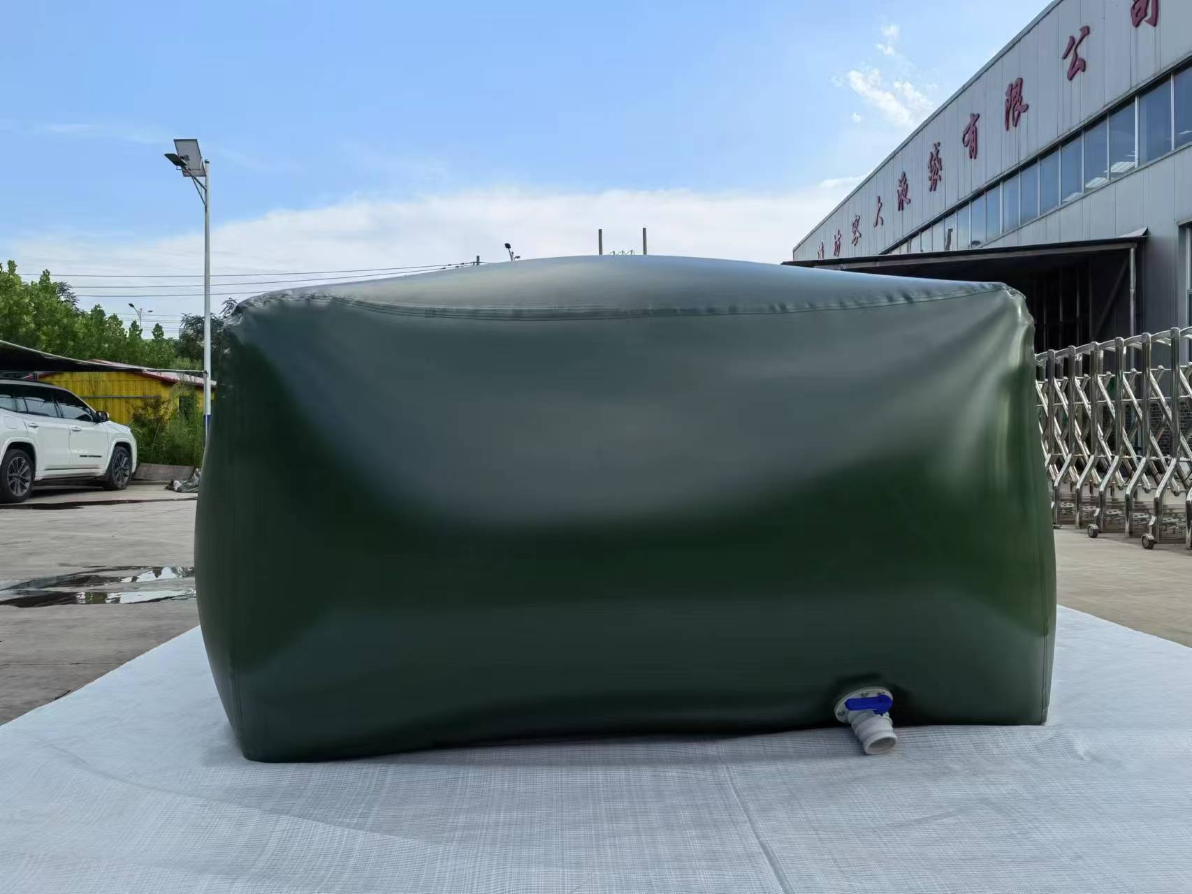 A large green inflatable water bladder with a white valve, resting on a white tarp on a concrete surface, is positioned in a parking lot with a white car, trees, and a building with red Chinese characters in the background under a partly cloudy sky.