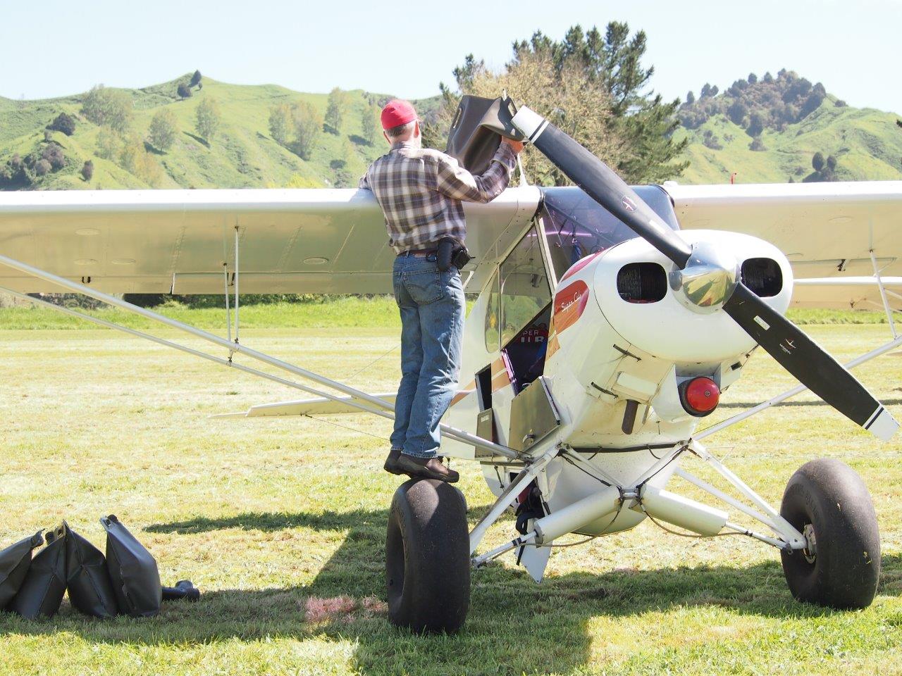 A man wearing a red cap, plaid shirt, and jeans stands on the wheel of a small white propeller airplane, refueling it with a flexible fuel storage bag in a grassy field with rolling green hills in the background.