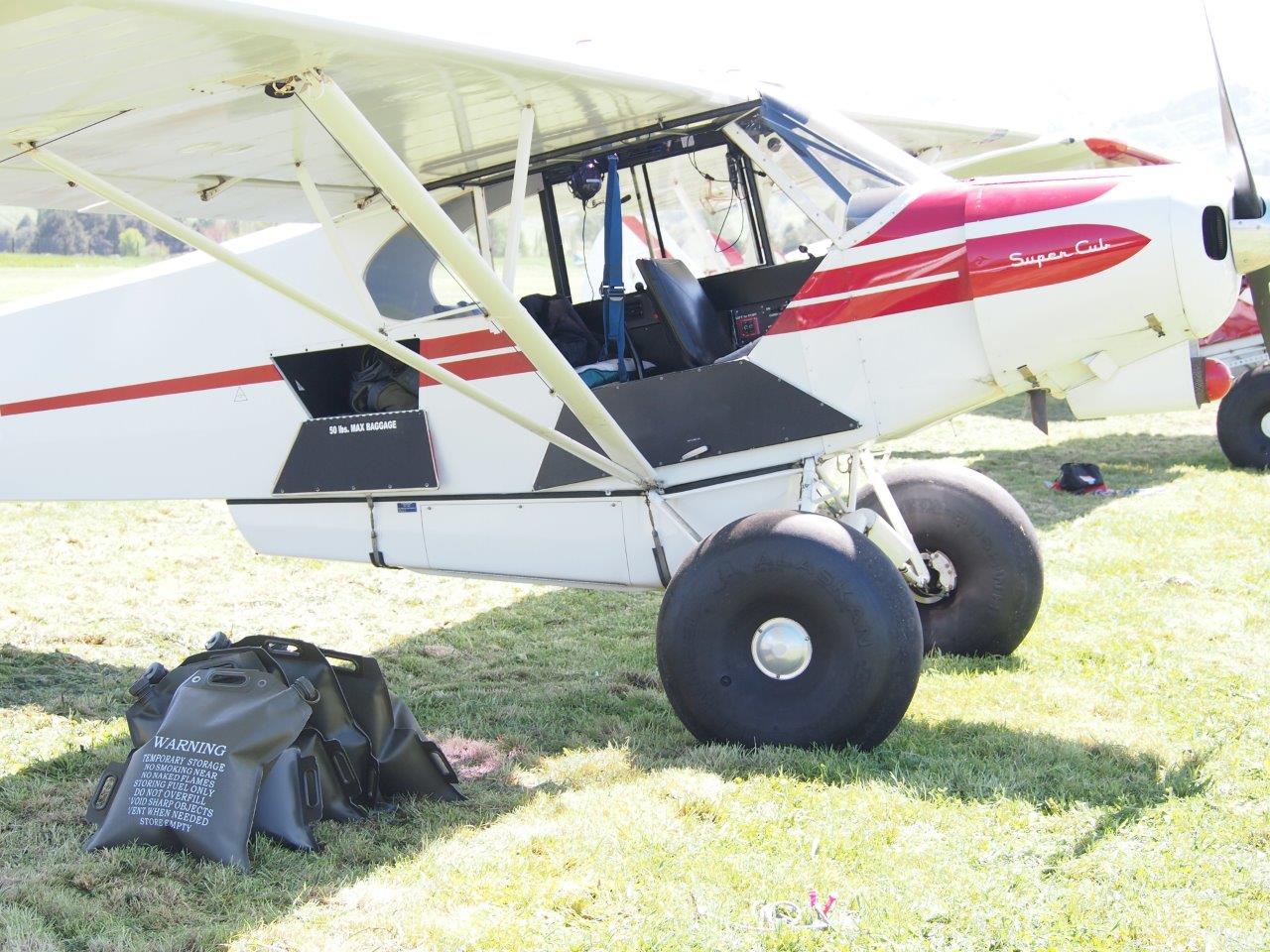 A white and red Super Cub aircraft is parked on a grassy field, with its cockpit open. A flexible fuel storage bag with a warning label is lying on the ground beside the plane. The background features a bright outdoor setting with green hills.