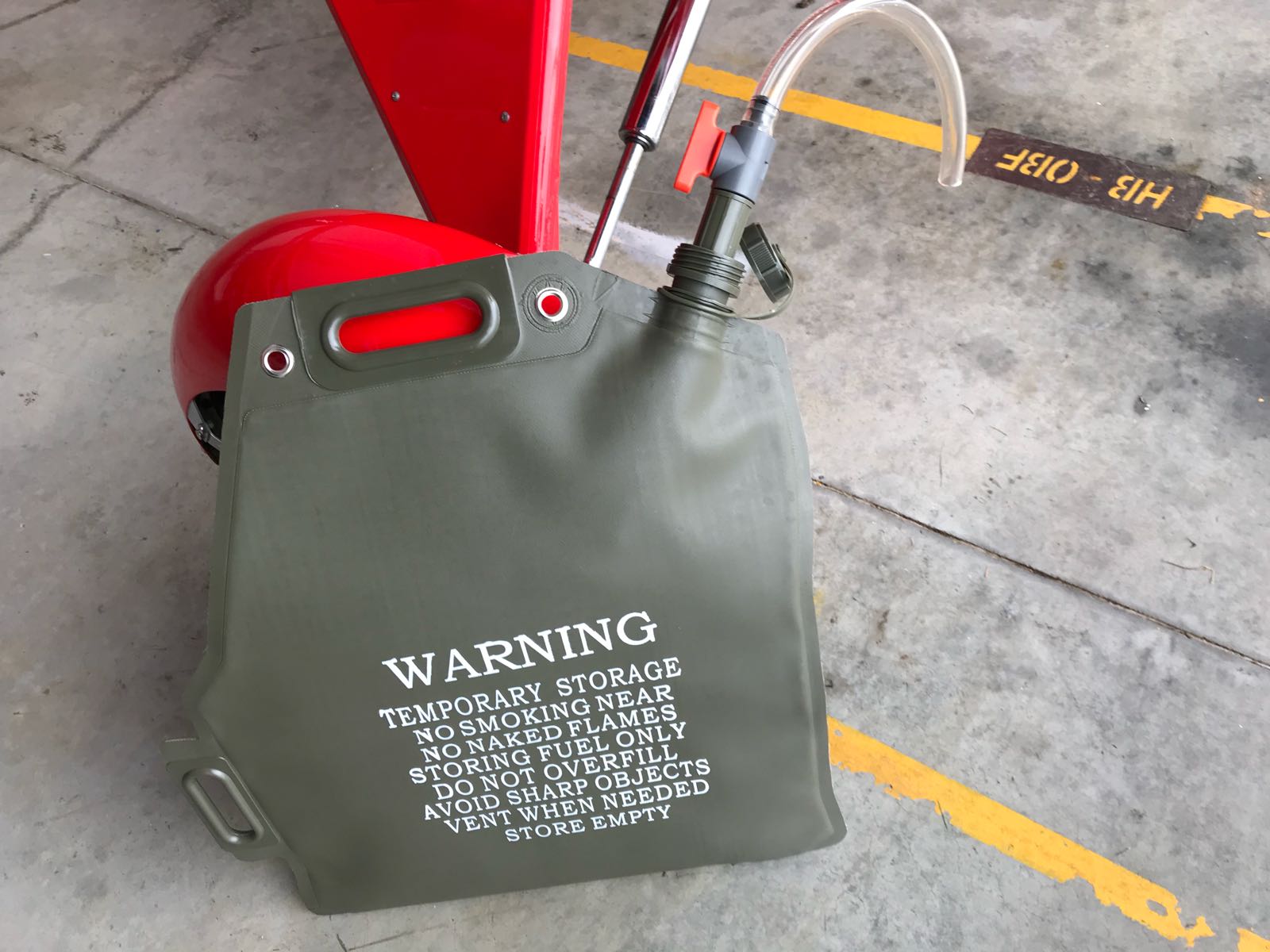 Portable military-style fuel storage bag with a built-in handle, nozzle, and warning instructions printed on the front, placed on a concrete floor near a red vehicle