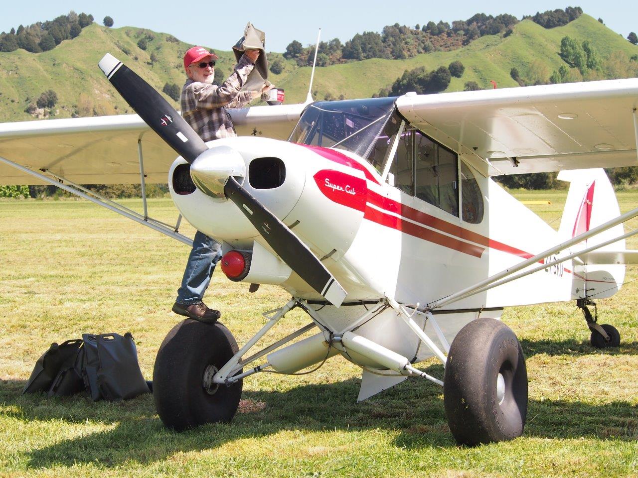This image shows a white and red Super Cub aircraft parked on a grassy field, with a man standing on the wheel to refuel or perform maintenance. He is wearing a plaid shirt, jeans, a red cap, and sunglasses. The background features green hills and a clear sky. There are flexible fuel storage bags on the ground beside the aircraft.